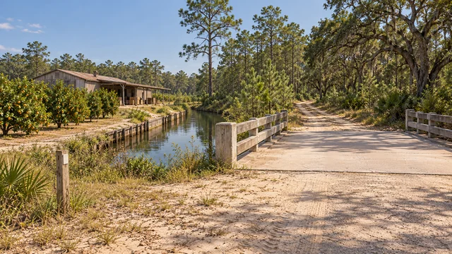 A low concrete crossing over a still canal, with a wooded path and modest building beyond.